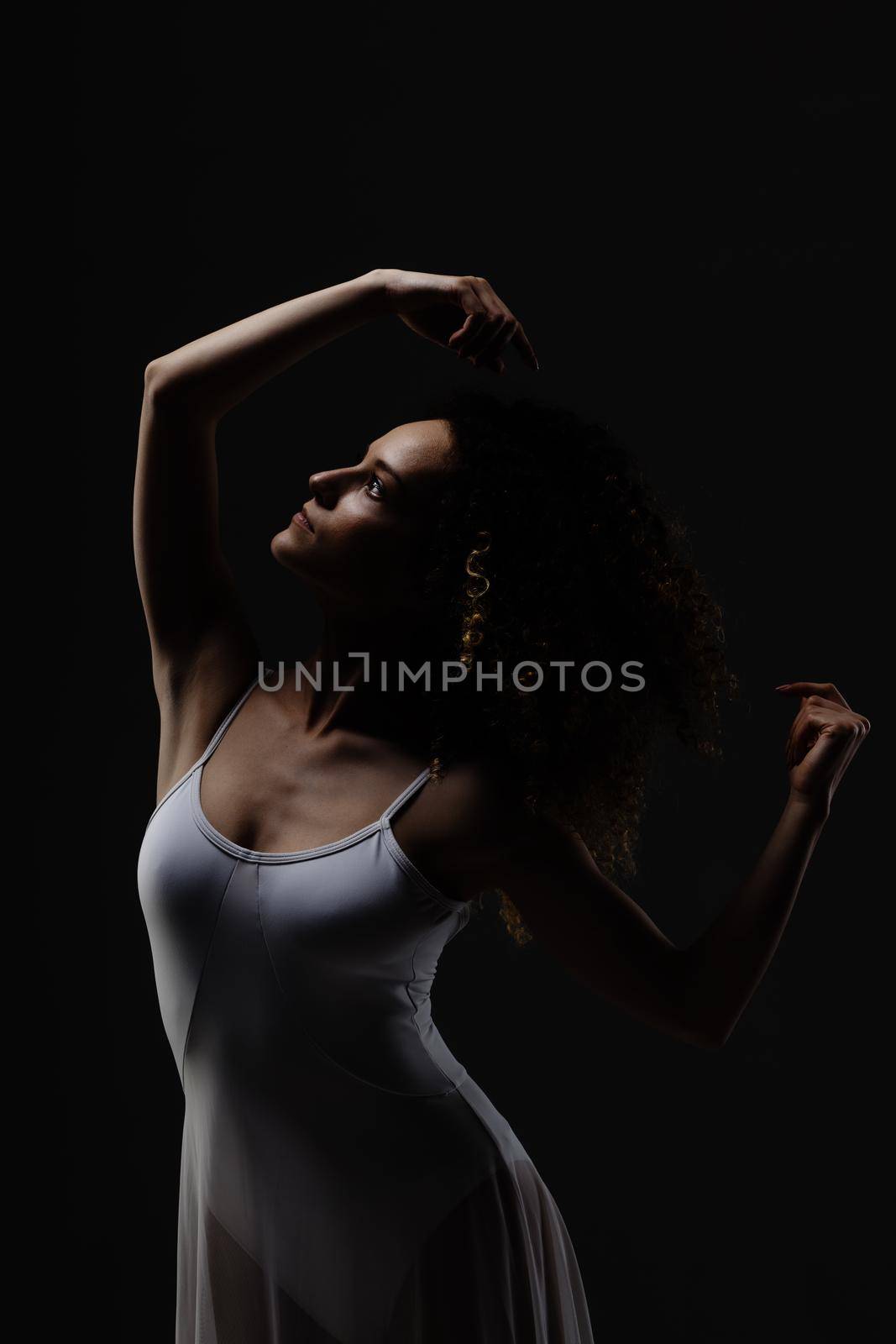 Girl with curly hair making ballet poses. Side lit silhouette of ballerina in white dress against black background.