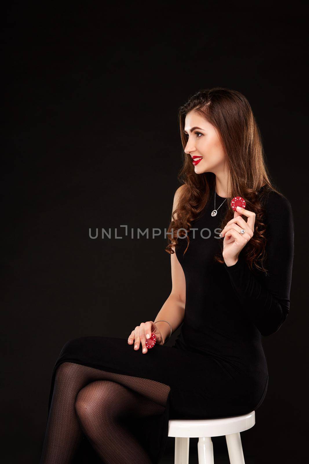 Sexy curly hair brunette in black dress posing with chips in her hands, poker concept black background. sits on a chair