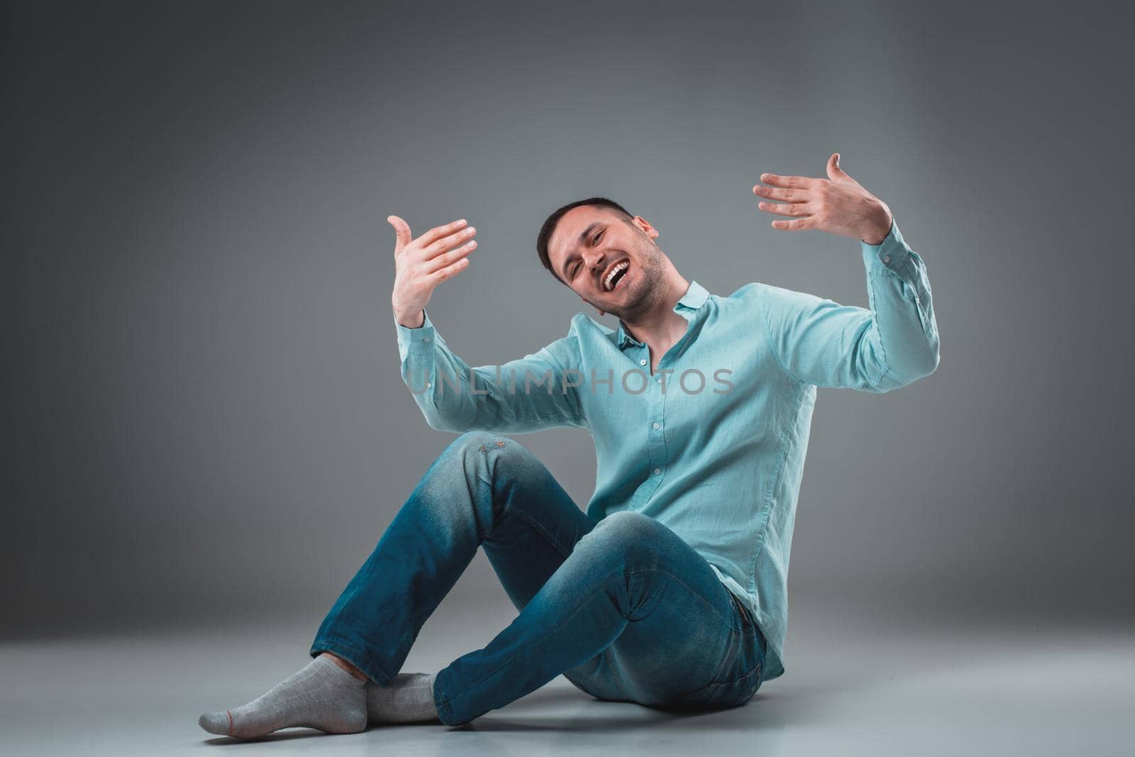 Appealing casual young man sitting on the floor, looking to the camera smiling. Studio shot.