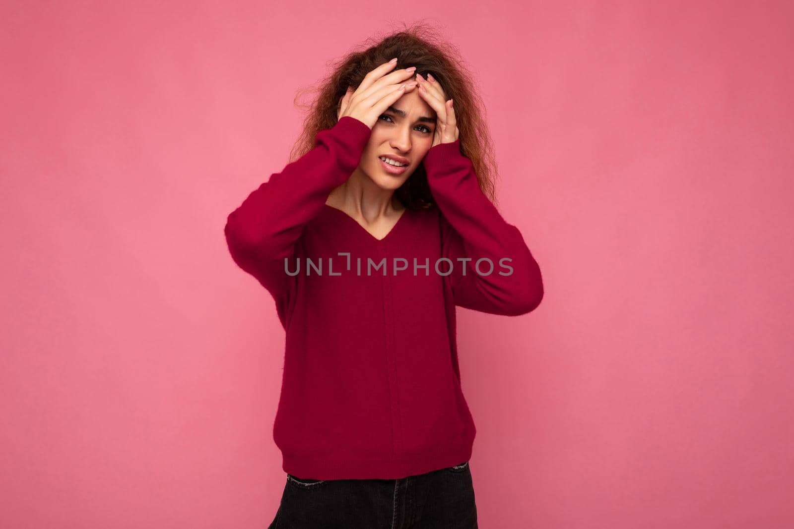 Portrait of young emotional beautiful brunette curly woman with sincere emotions wearing trendy pink pullover isolated on pink background with copy space and having headache.