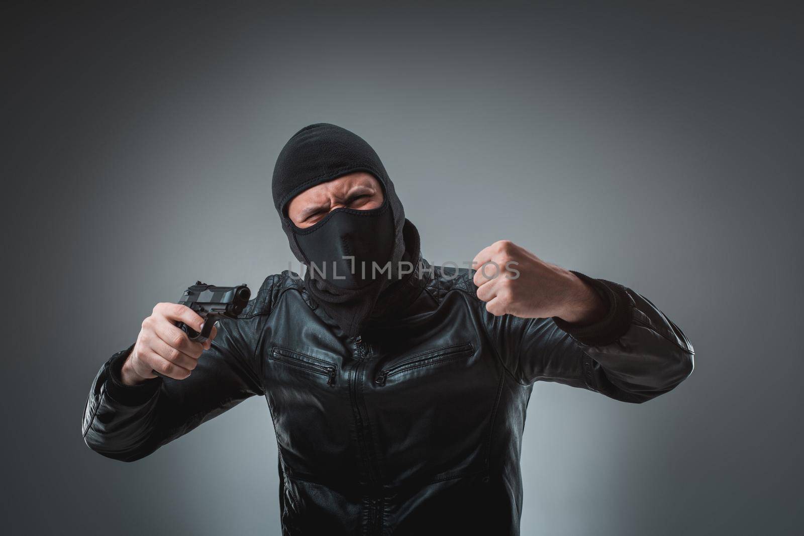 Masked robber with gun, looking into the camera. Studio shot on gray background. Emotions