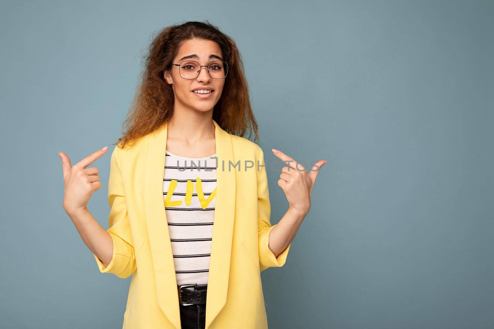 Photo of young positive beautiful brunette curly woman with sincere emotions wearing casual yellow jacket and optical glasses isolated on blue background with copy space and asking pointing at yourself.