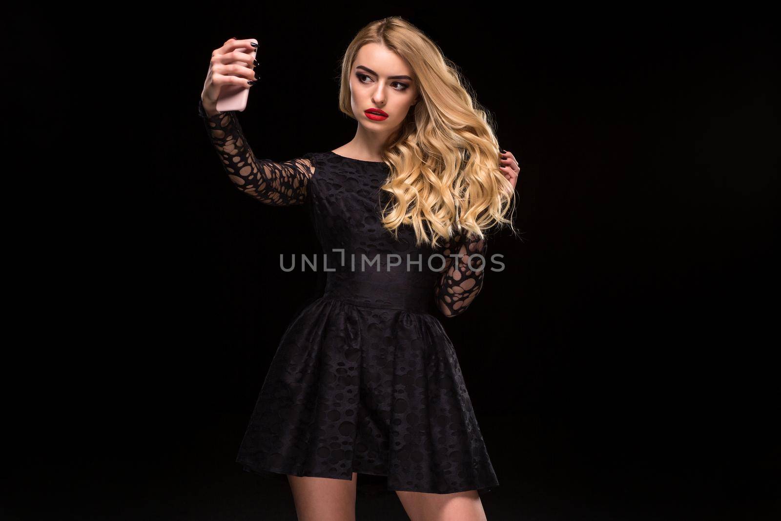 Beautiful blonde woman in black dress and make up taking selfie with mobile phone isolated on the black background. Female beauty model with curly hair.