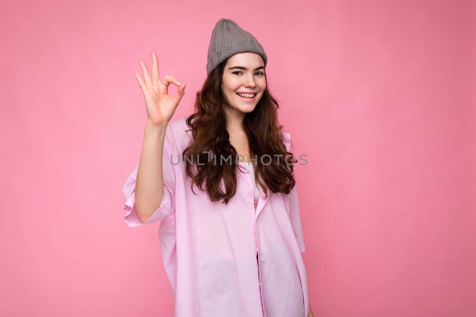 Photo of young positive happy smiling beautiful woman with sincere emotions wearing stylish clothes isolated over background with copy space and showing ok gesture. It's okay.