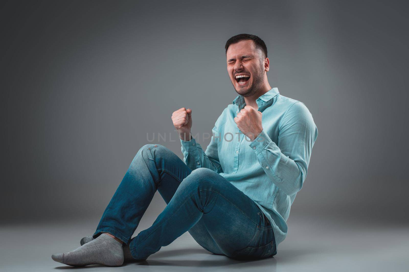 The man is sitting on the floor, isolated on gray background. Man showing different emotions. A man is dressed in blue jeans and a shirt