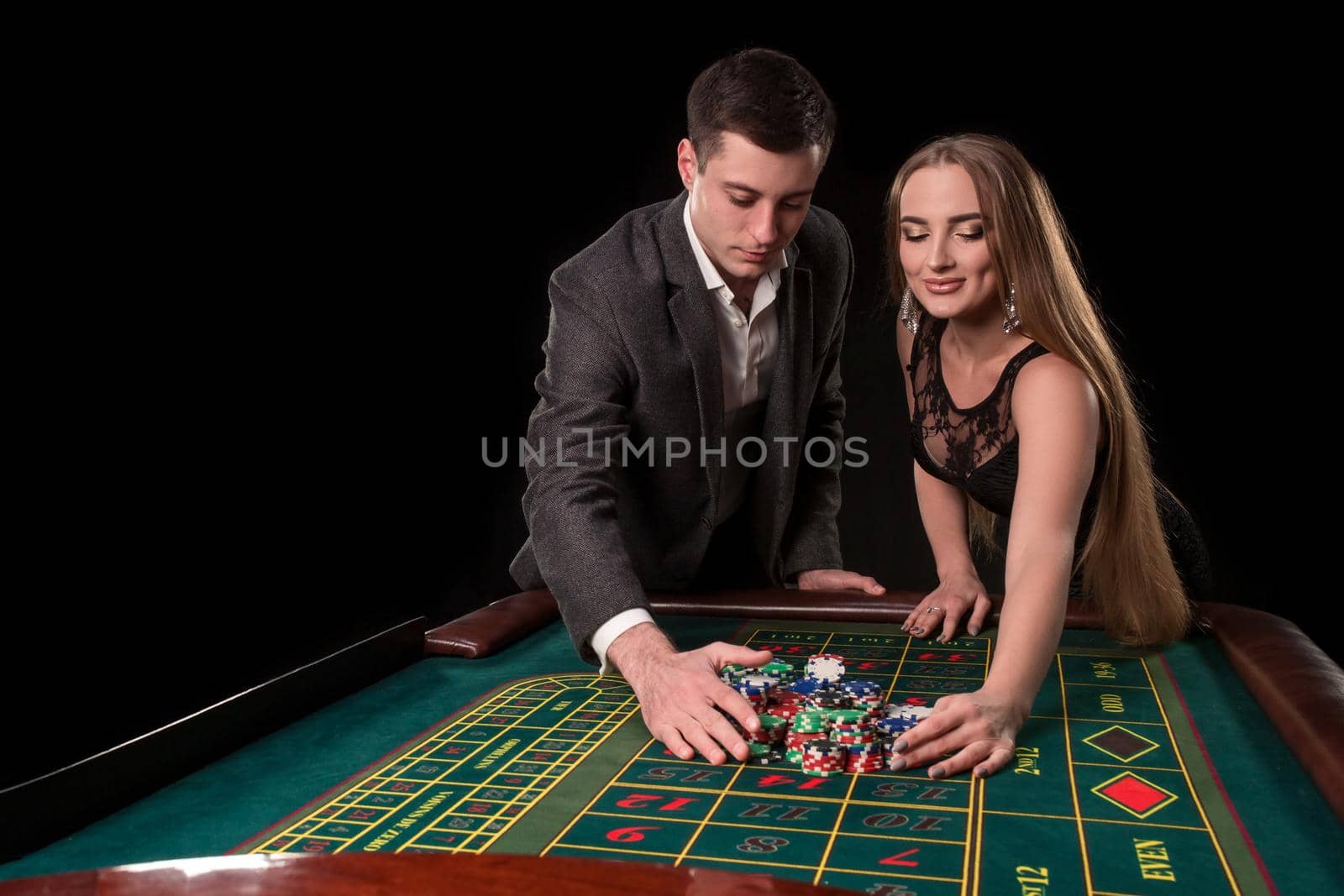 Young beautiful couple takes their winnings at the roulette table at the casino, on a black background. A man in a suit with a woman in a black dress