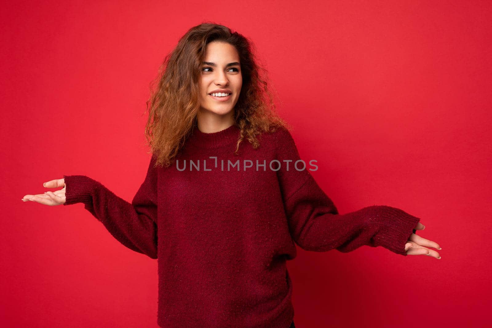 Adult stylish pretty curly brunette female person with sincere emotions poising isolated on background wall with copy space wearing casual dark red sweater. Positive concept.