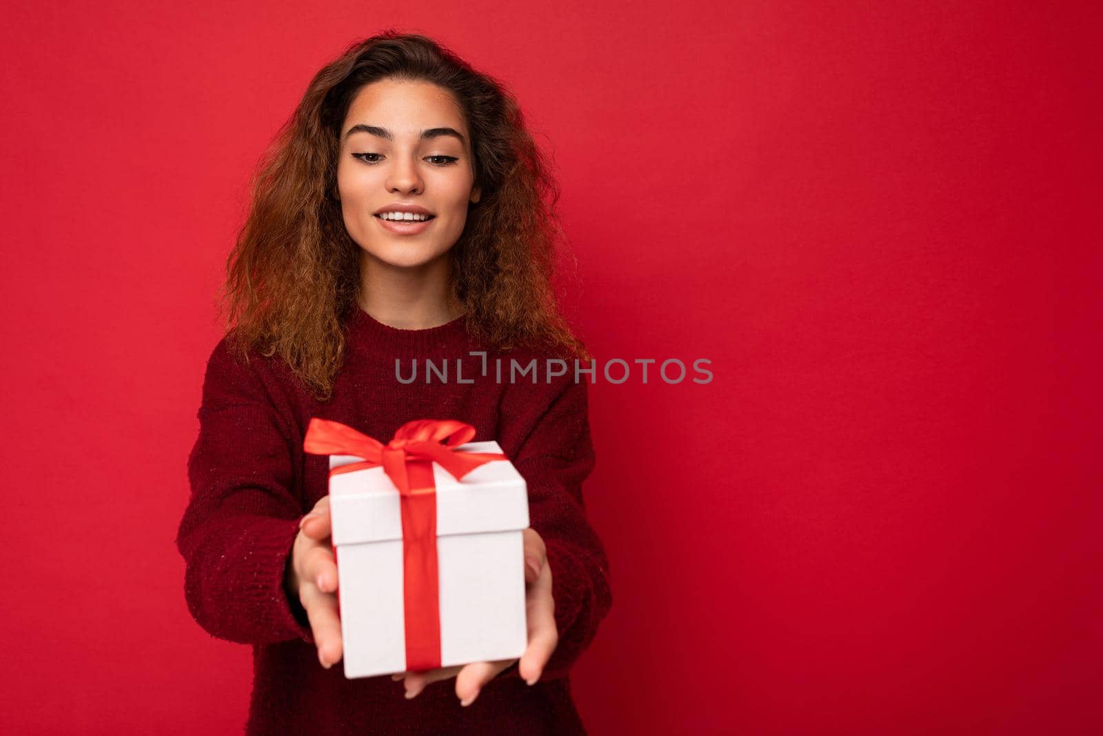 Attractive positive smiling young brunette curly woman isolated over red background wall wearing red sweater holding gift box looking at present white box with red ribbon. Empty space