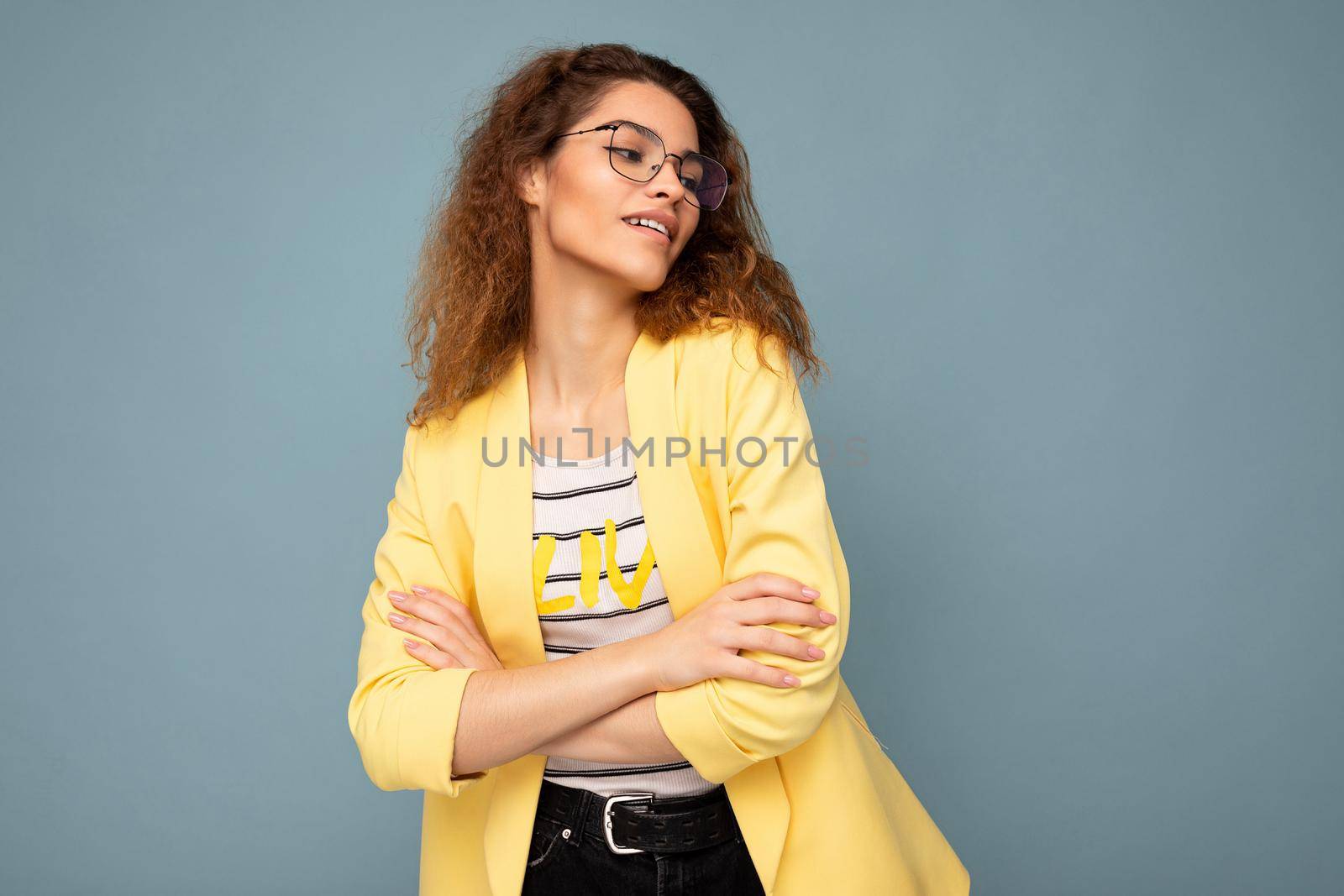 Photo of young positive arrogant beautiful brunette curly woman with sincere emotions wearing casual yellow jacket and optical glasses isolated on blue background with copy space.