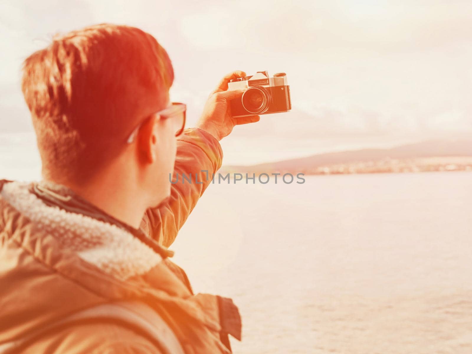 Traveler young man doing selfie with old photo camera on coastline. Image with sunlight effect. Focus on camera