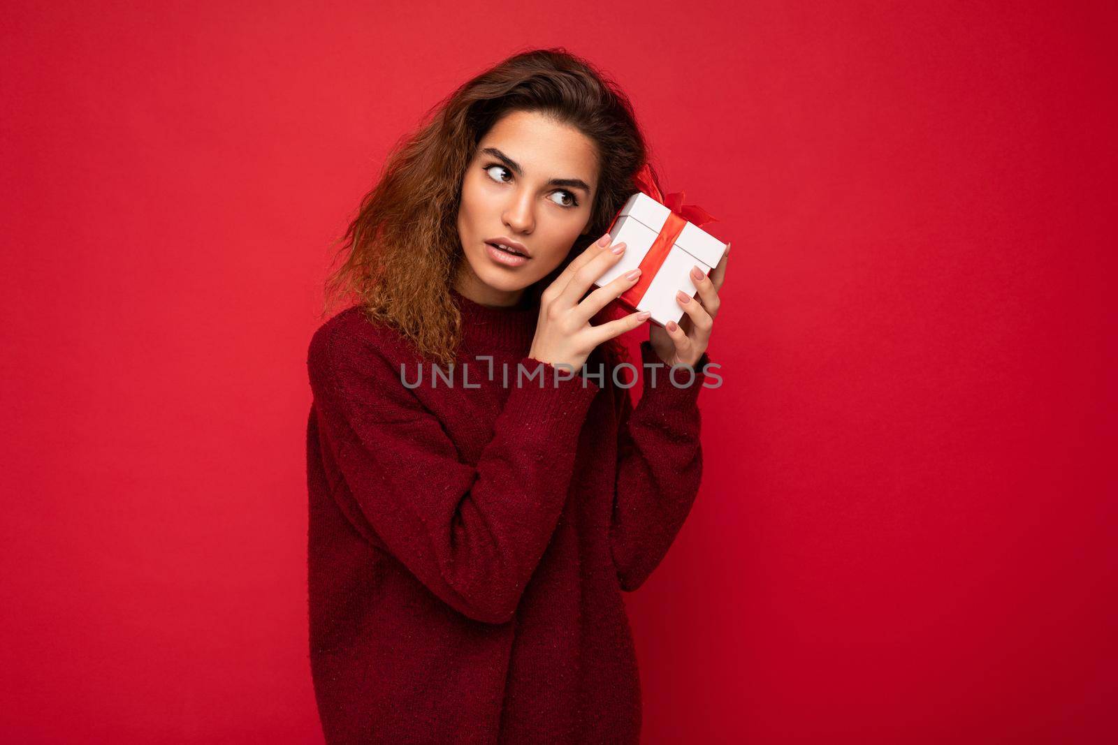 Photo shot of attractive serious thoughtful concentrated young brunette curly woman isolated over red background wall wearing red sweater holding gift box looking to the side and thinking.