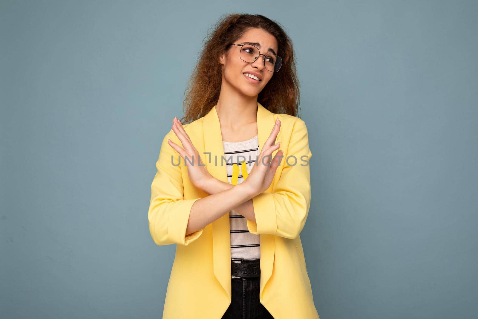 Photo of young emotional attractive brunette curly woman with sincere emotions wearing stylish yellow jacket and optical glasses isolated on blue background with empty space and saying I don't want it.