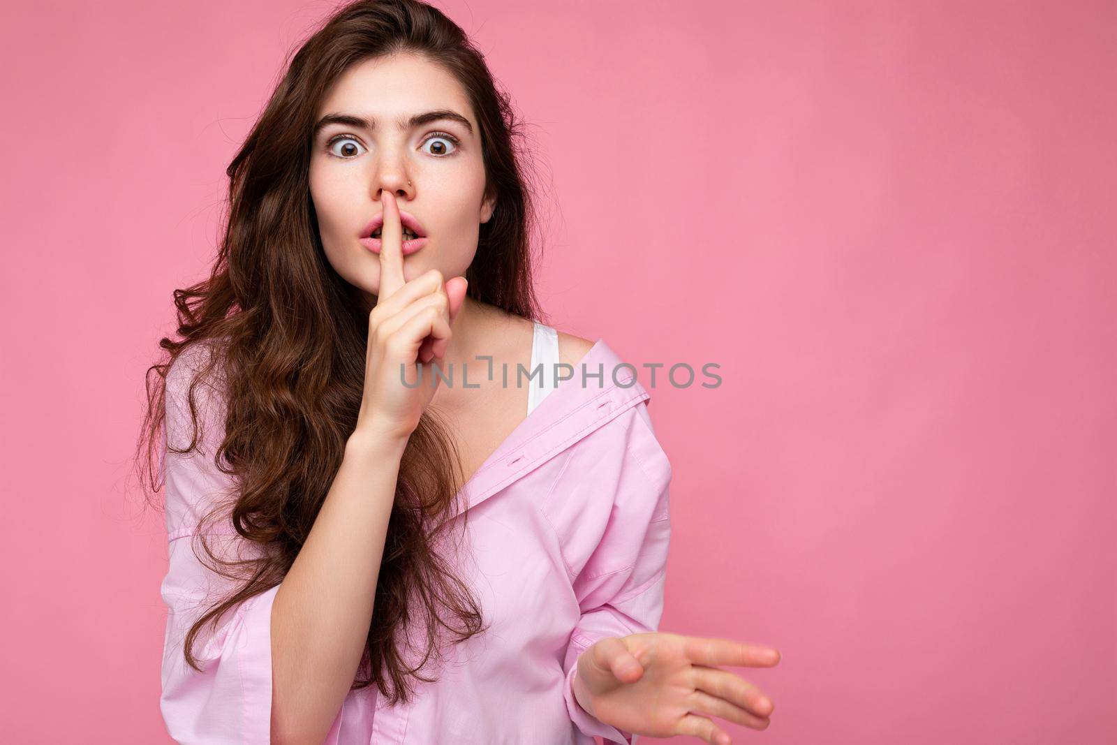 Photo of young shocked beautiful wavy-haired brunette woman with sincere emotions wearing casual pink shirt isolated over pink background with copy space and showing silence gesture with finger near lips. Shhh sign.
