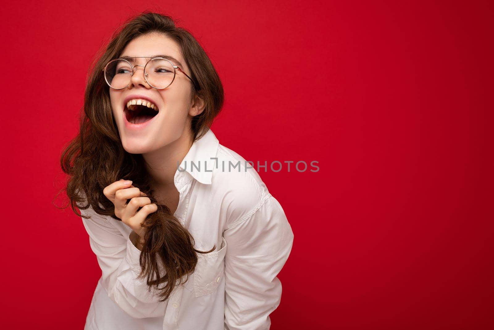 Beautiful young happy joyful funny curly brunette woman wearing white shirt and optical glasses isolated on red background with copy space. Emotional concept