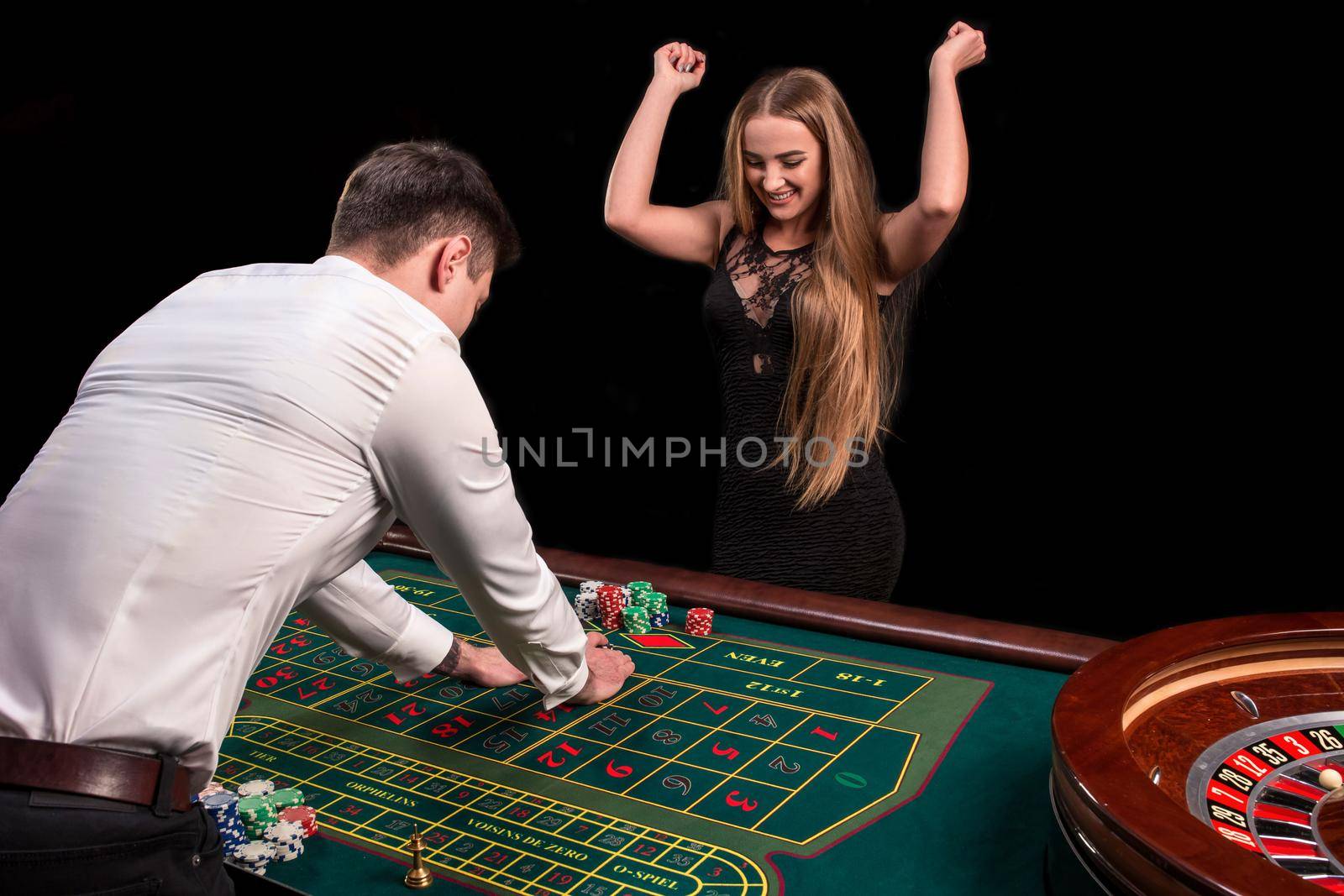 A close-up on the back of the croupier in a white shirt, image of green casino table with roulette and chips, a rich woman betting of gambling in the background by nazarovsergey