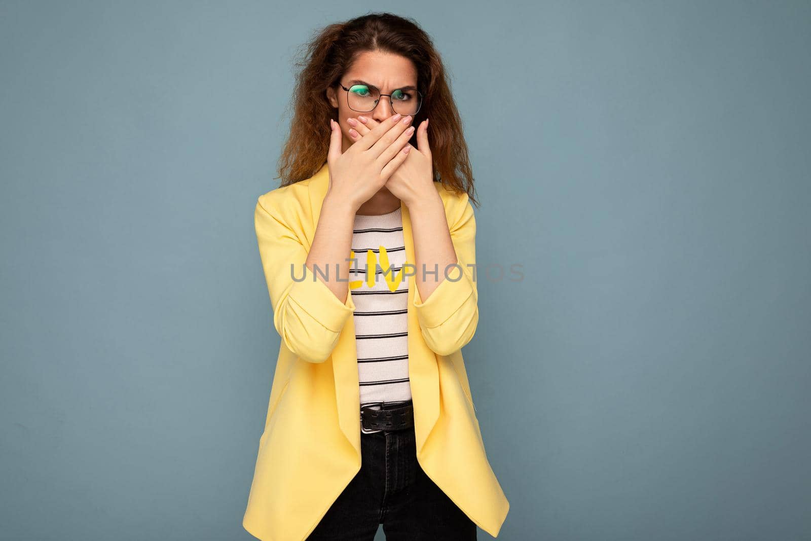 Photo shot of young positive happy nice cute brunette curly woman with sincere emotions wearing trendy yellow jacket isolated on blue background with copy space and covering mouth.