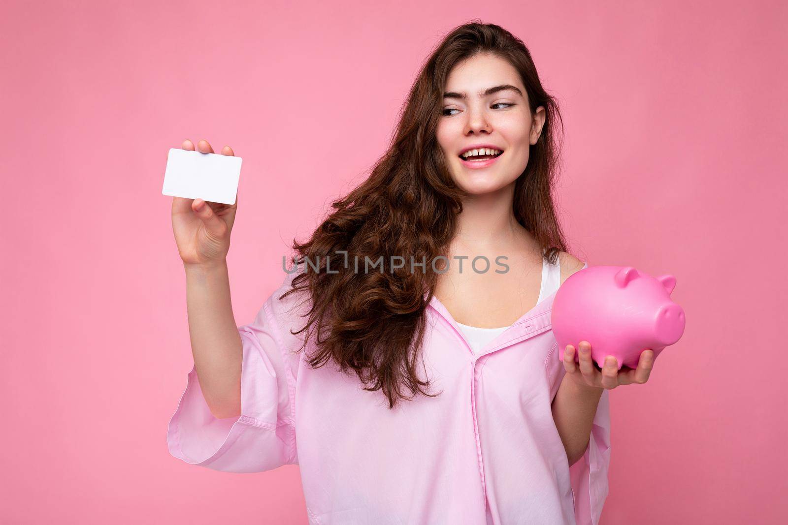Portrait of beautiful positive cheerful cute smiling young brunette woman in stylish shirt isolated on pink background with copy space and holding pink pig moneybox and credit card for mockup.