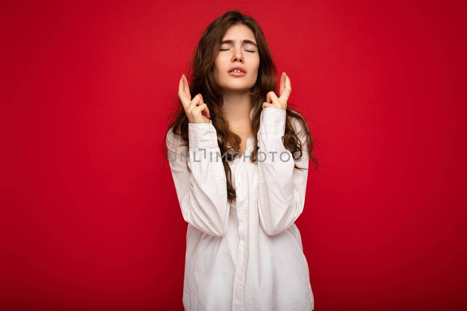 Shot of attractive young curly brunette woman wearing white shirt isolated on red background with copy space, praying and dreaming.