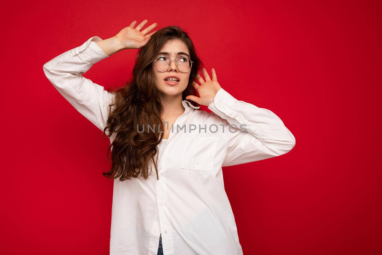 Shot of attractive angry sad dissatisfied young curly brunette female person wearing white shirt and optical glasses isolated on red background with free space.