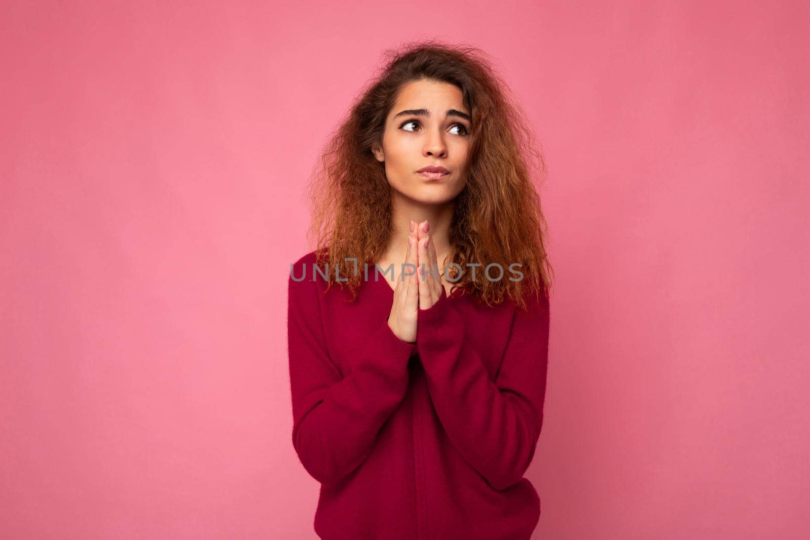 Photo of young emotional beautiful brunette curly woman with sincere emotions wearing trendy pink pullover isolated over pink background with free space and keeping hands in praying gesture and believing in good luck.