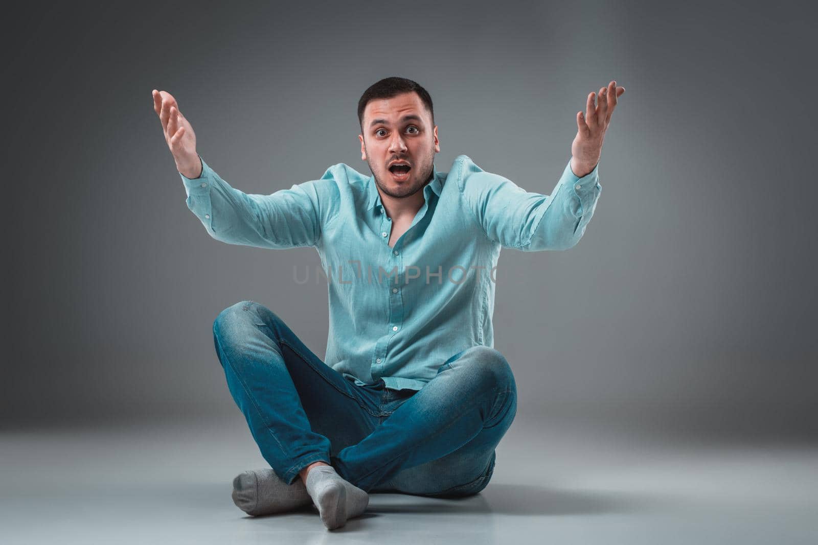 Handsome young man sitting on a floor with raised hands, isolated on gray background. Emotion concept