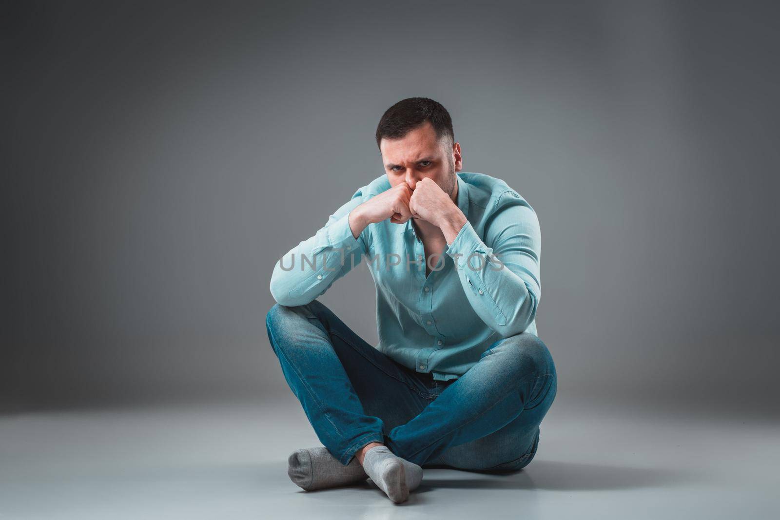The man is sitting on the floor, isolated on gray background. Man showing different emotions. A man is dressed in blue jeans and a shirt