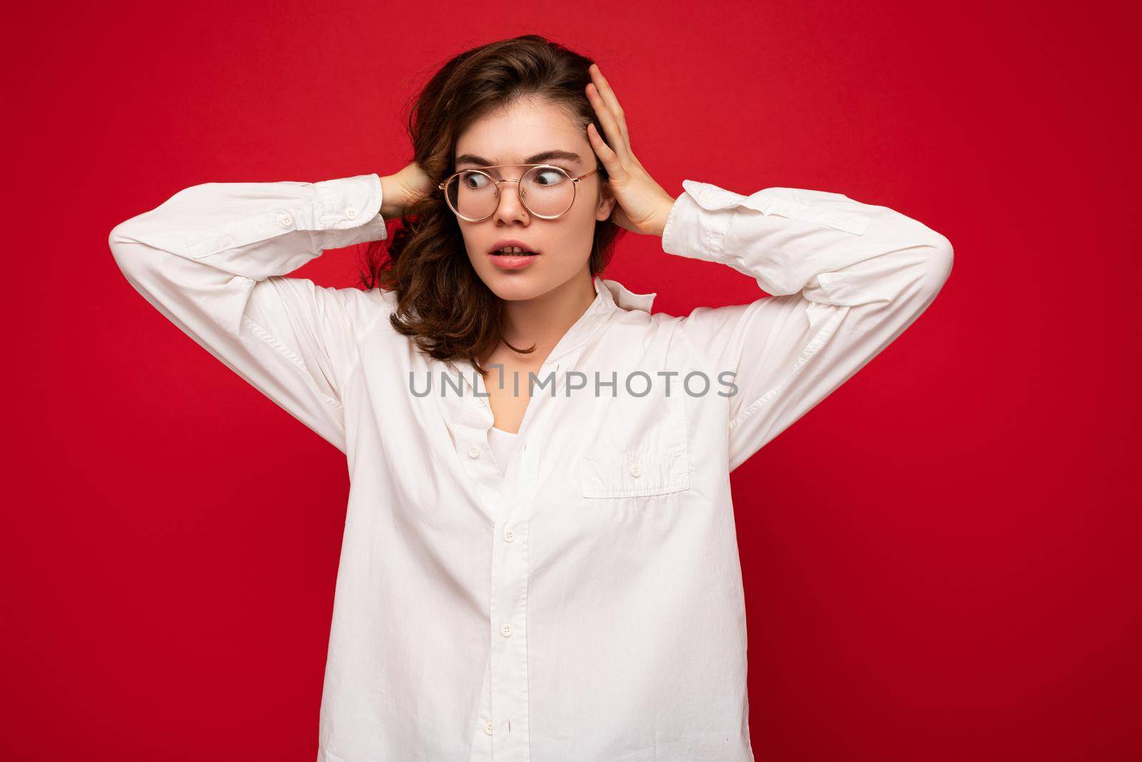 Beautiful young amazed curly brunette woman wearing white shirt and optical glasses isolated on red background with copy space. Shock concept.