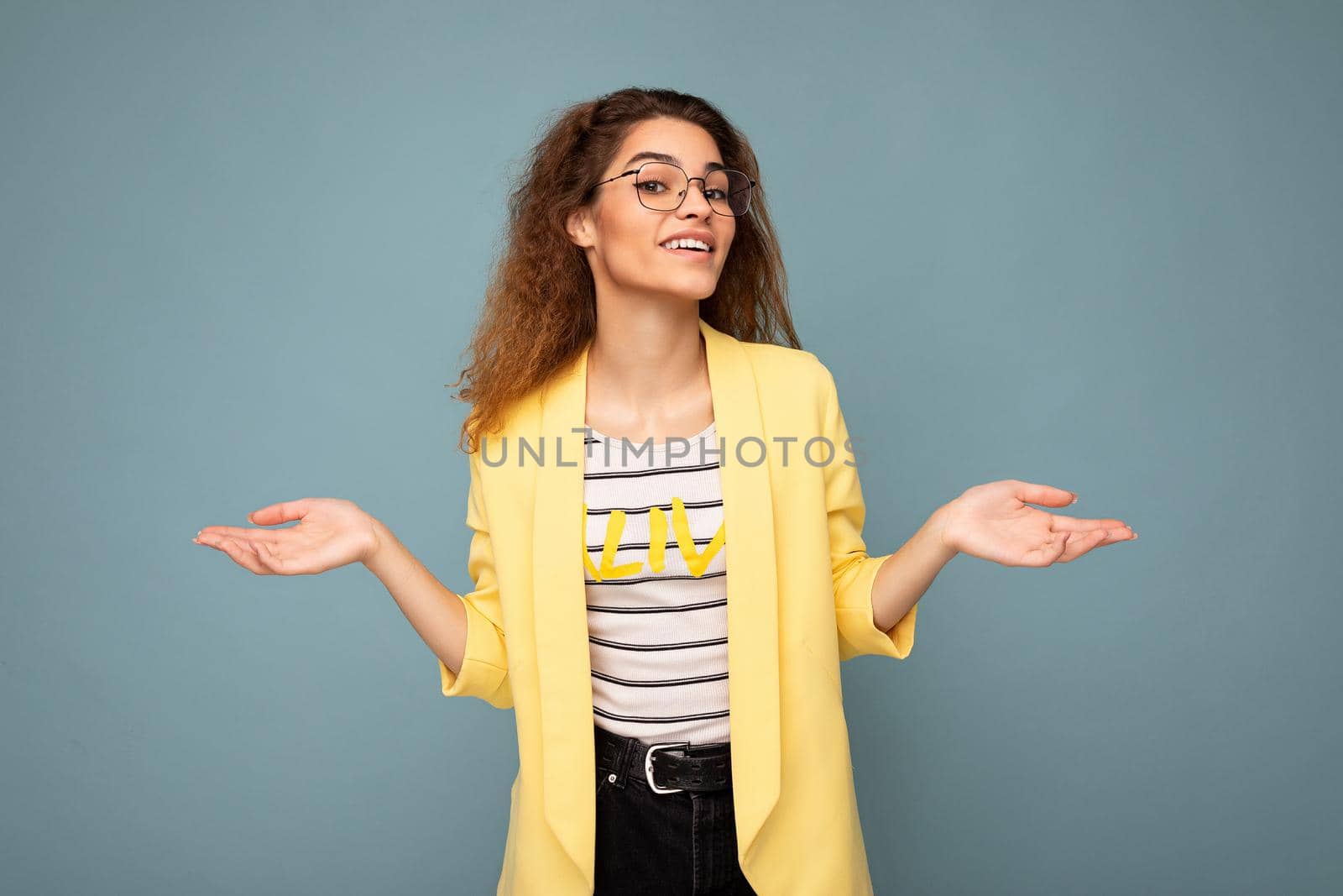 Photo of young positive thoughtful beautiful brunette curly woman with sincere emotions wearing casual yellow jacket and optical glasses isolated on blue background with copy space and having doubts.