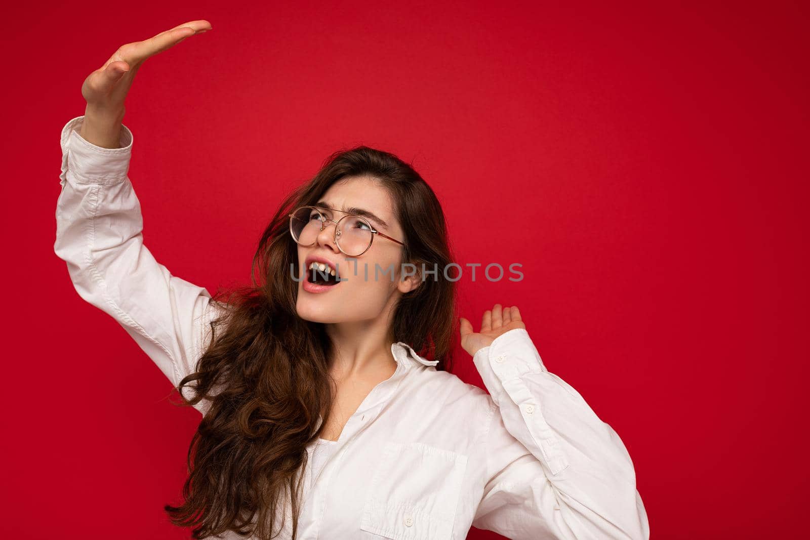 Shot of attractive dissatified young curly brunette woman wearing white shirt and optical glasses isolated on red background with free space.