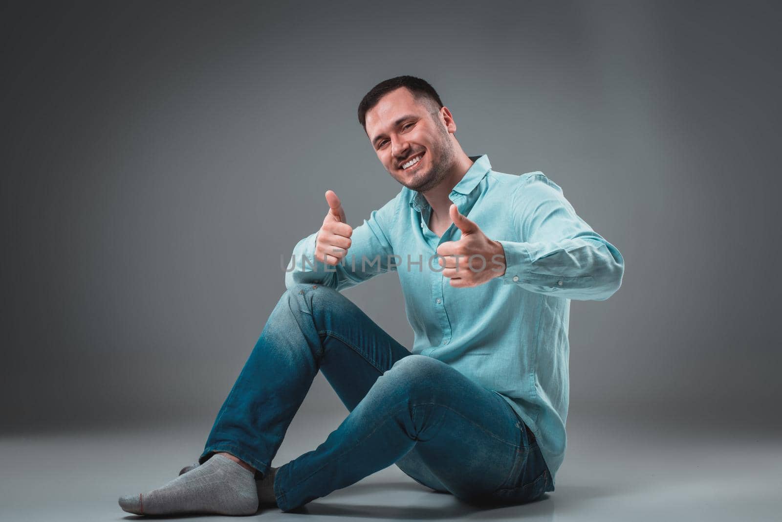 Handsome young man sitting on a floor with raised hands gesturing happiness on gray background. A man in jeans and a blue shirt