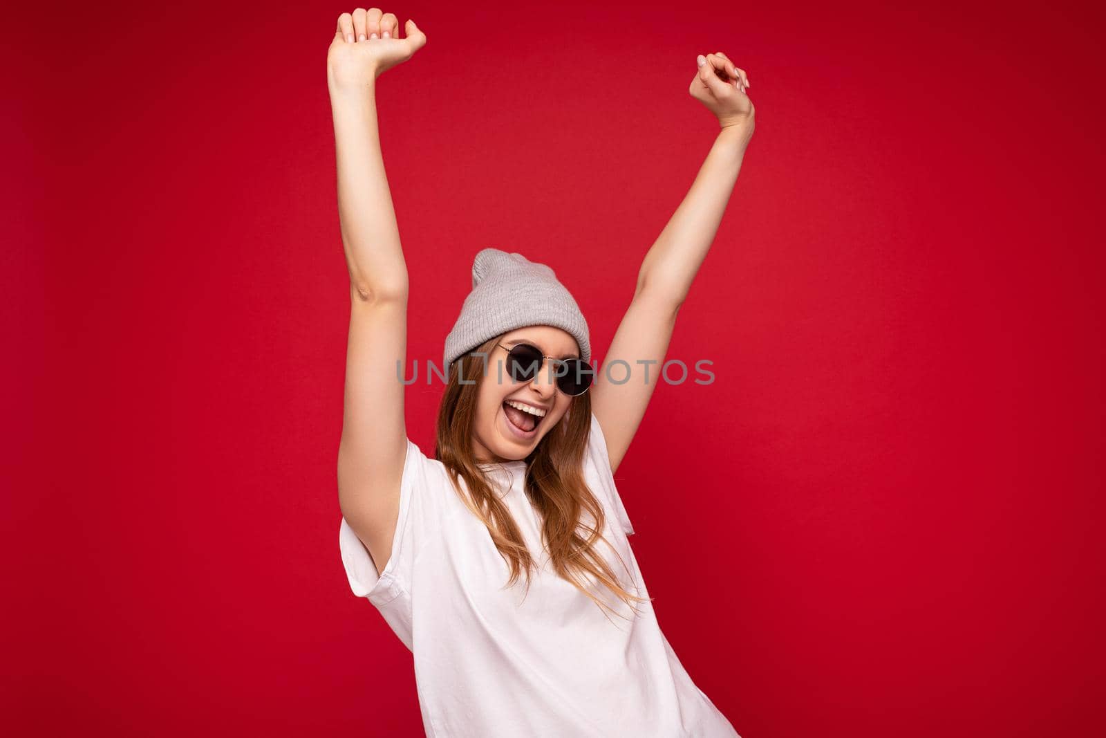 Photo of young emotional positive happy attractive dark blonde woman with sincere emotions wearing casual white t-shirt with empty space for mockup gray hat and sunglasses isolated on red background with copy space and celebrating victory showing yeah gesture.