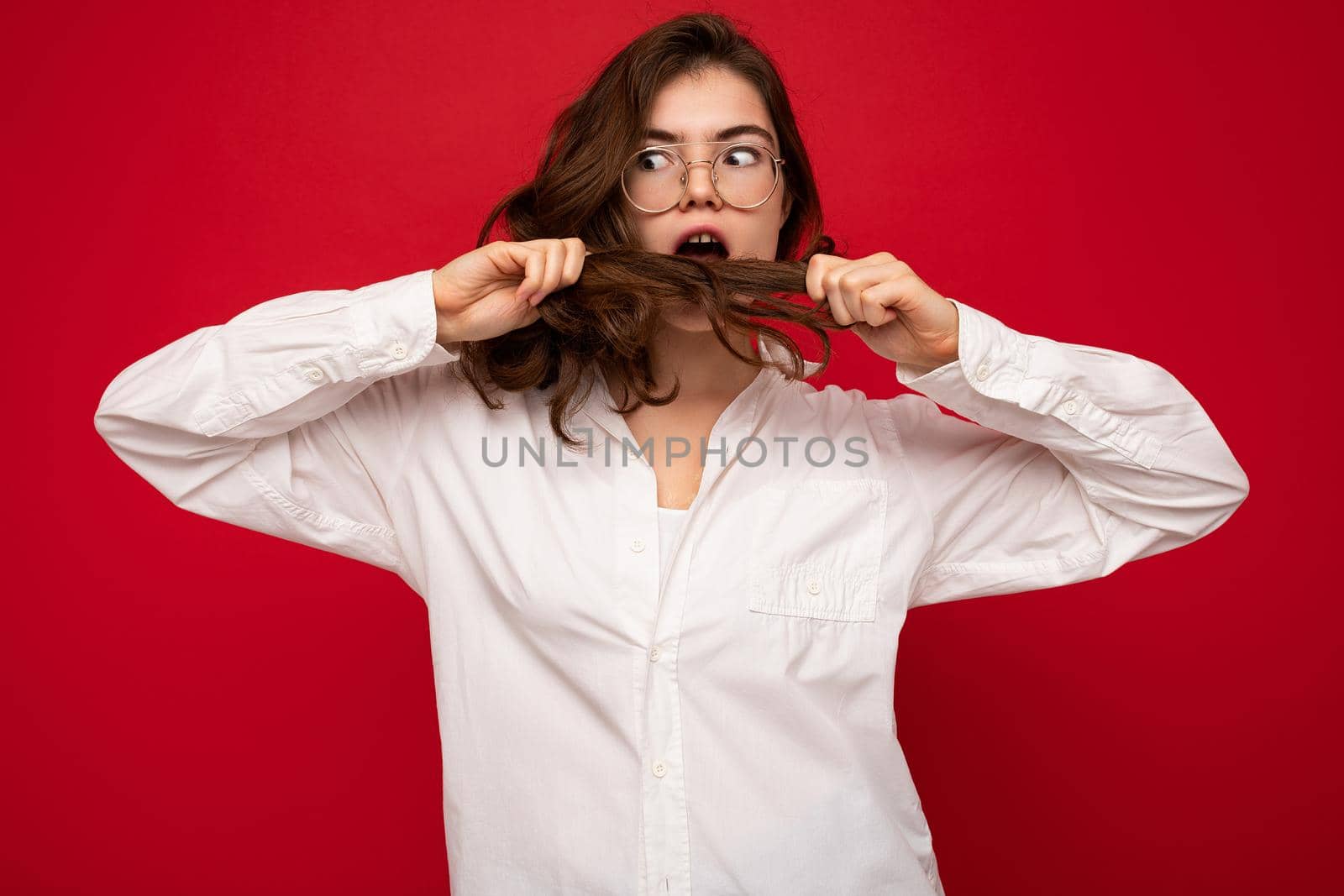 Beautiful young amazed shocked brunette woman wearing white shirt and optical glasses. Sexy carefree female person posing isolated on red background with copy space and touching brown hair.