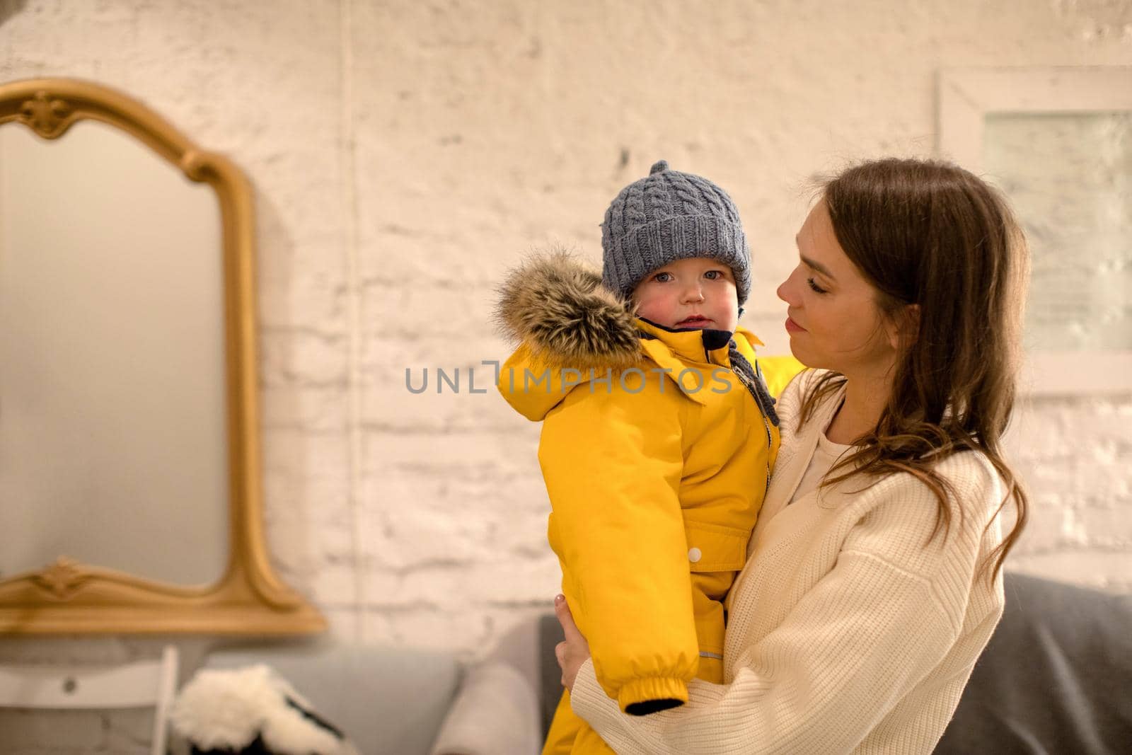 Little kid in yellow warm jacket and hat looking at camera in arms of young mother at home