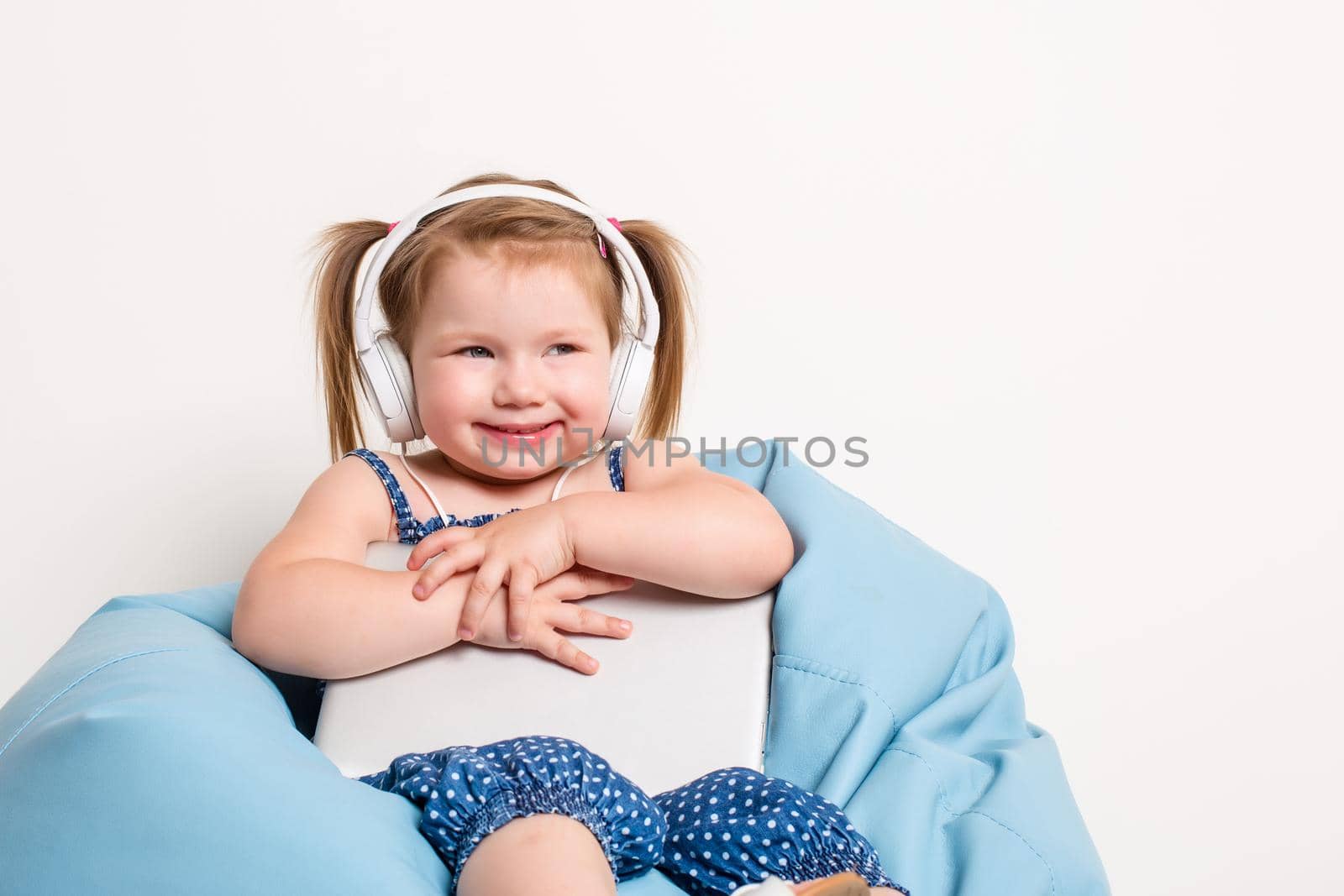 Cute little girl in headphones listening to music using a tablet and smiling while sitting on blue big bag. On white background. A child looks at the camera