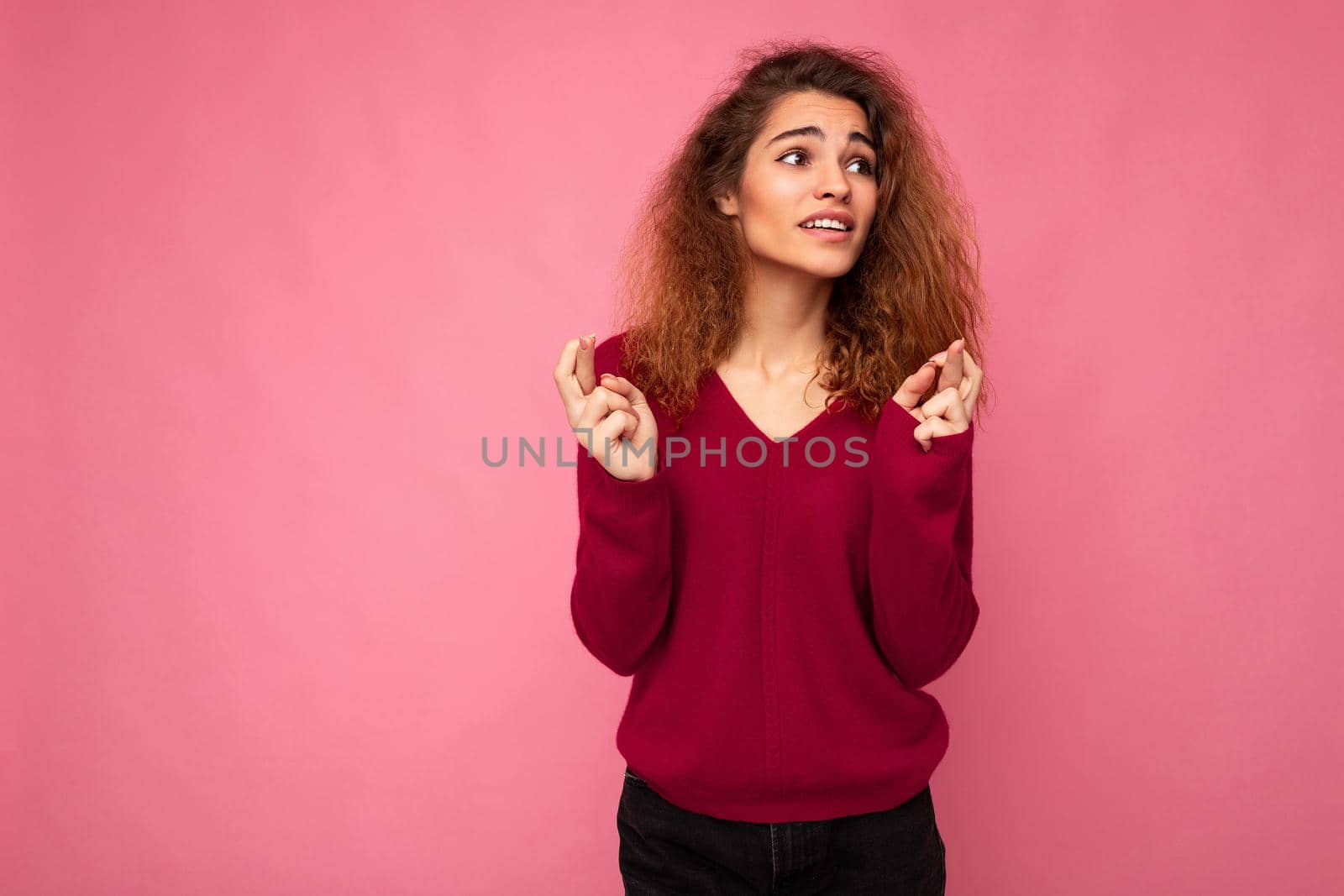 Photo of young emotional attractive brunette curly woman with sincere emotions wearing trendy pink sweater isolated over pink background with empty space and holding fingers crossed for good luck. Gesture concept.