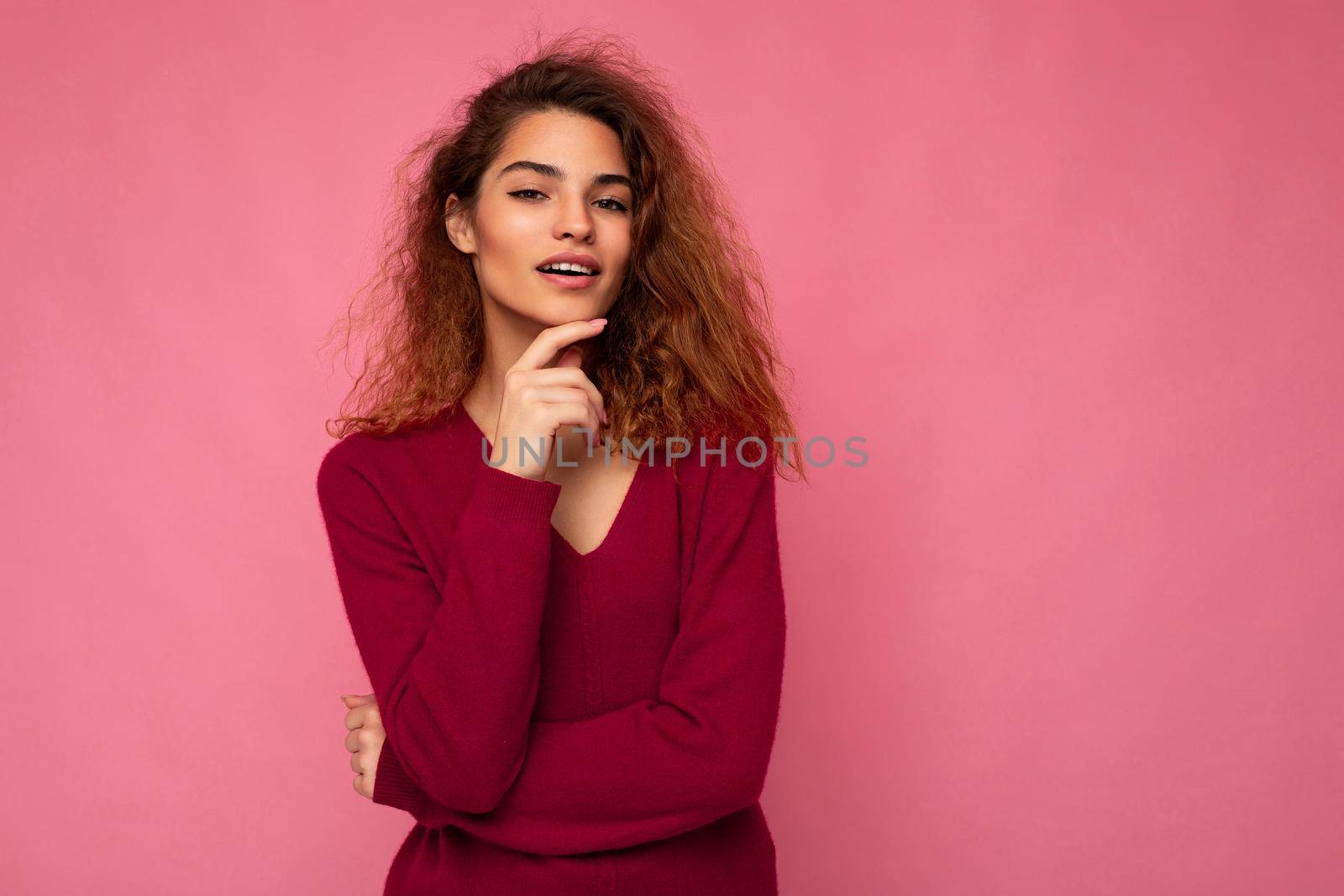 Portrait of young positive thoughtful beautiful brunette curly woman with sincere emotions wearing trendy pink pullover isolated on pink background with copy space.