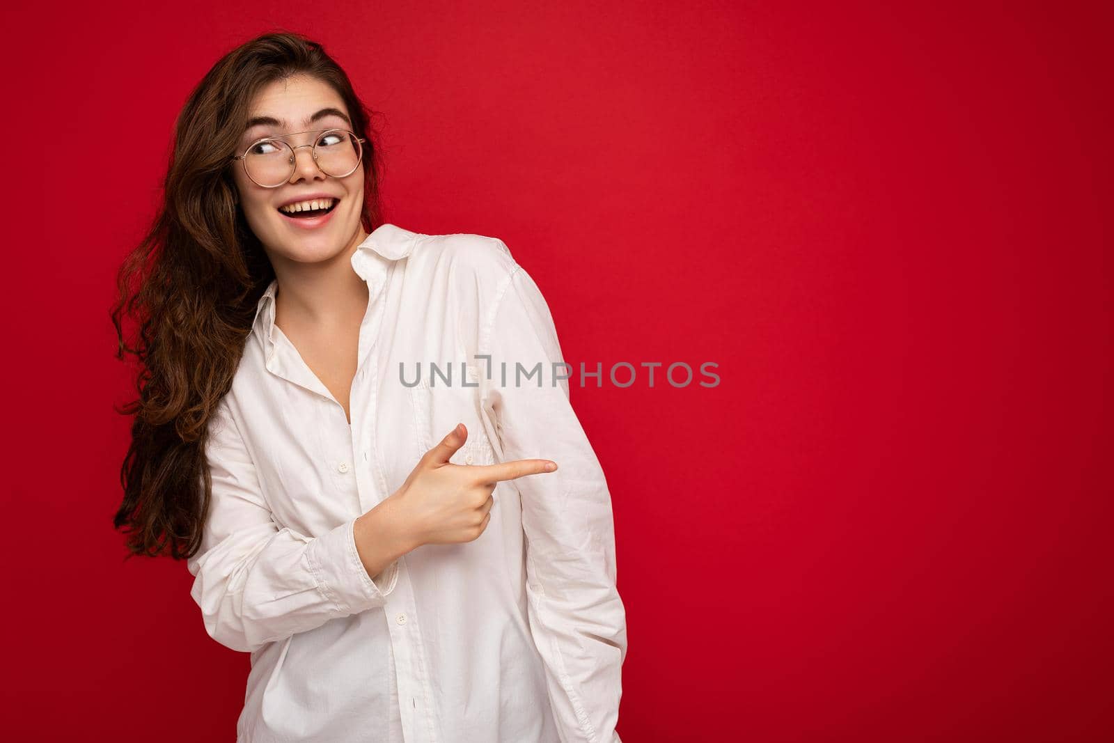 Photo of young beautiful happy smiling brunette woman wearing white shirt and optical glasses. Sexy carefree female person posing isolated near red wall in studio with free space. Positive model with natural makeup.