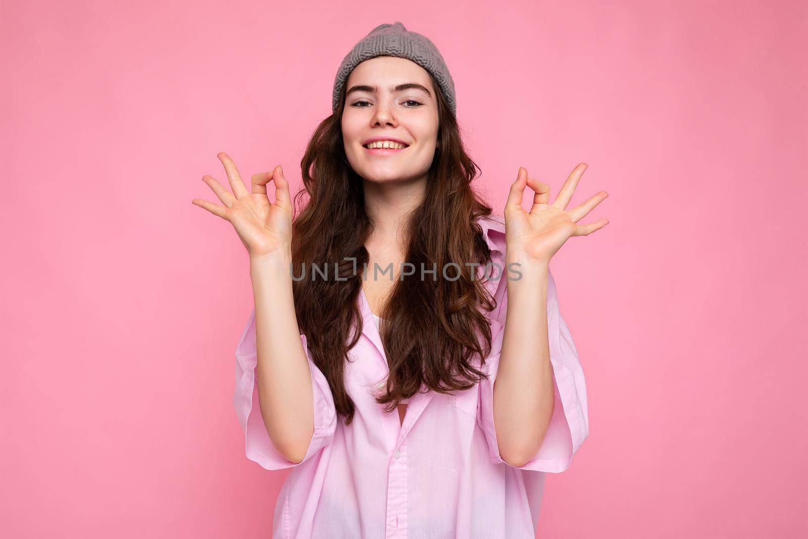 Portrait of beautiful positive cheerful cute smiling young brunette woman in stylish shirt and hipster grey hat isolated on pink background with copy space and showing okay gesture.
