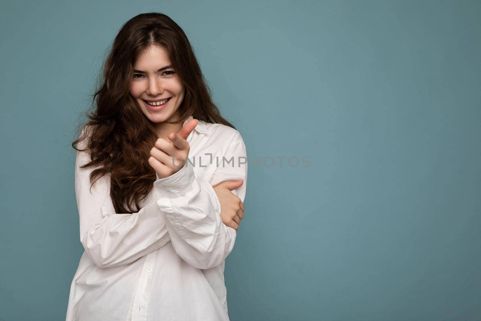 Portrait of young positive happy smiling cite nice curly brunette woman with sincere emotions wearing casual white shirt isolated on blue background with empty space and pointing at camera by TRMK