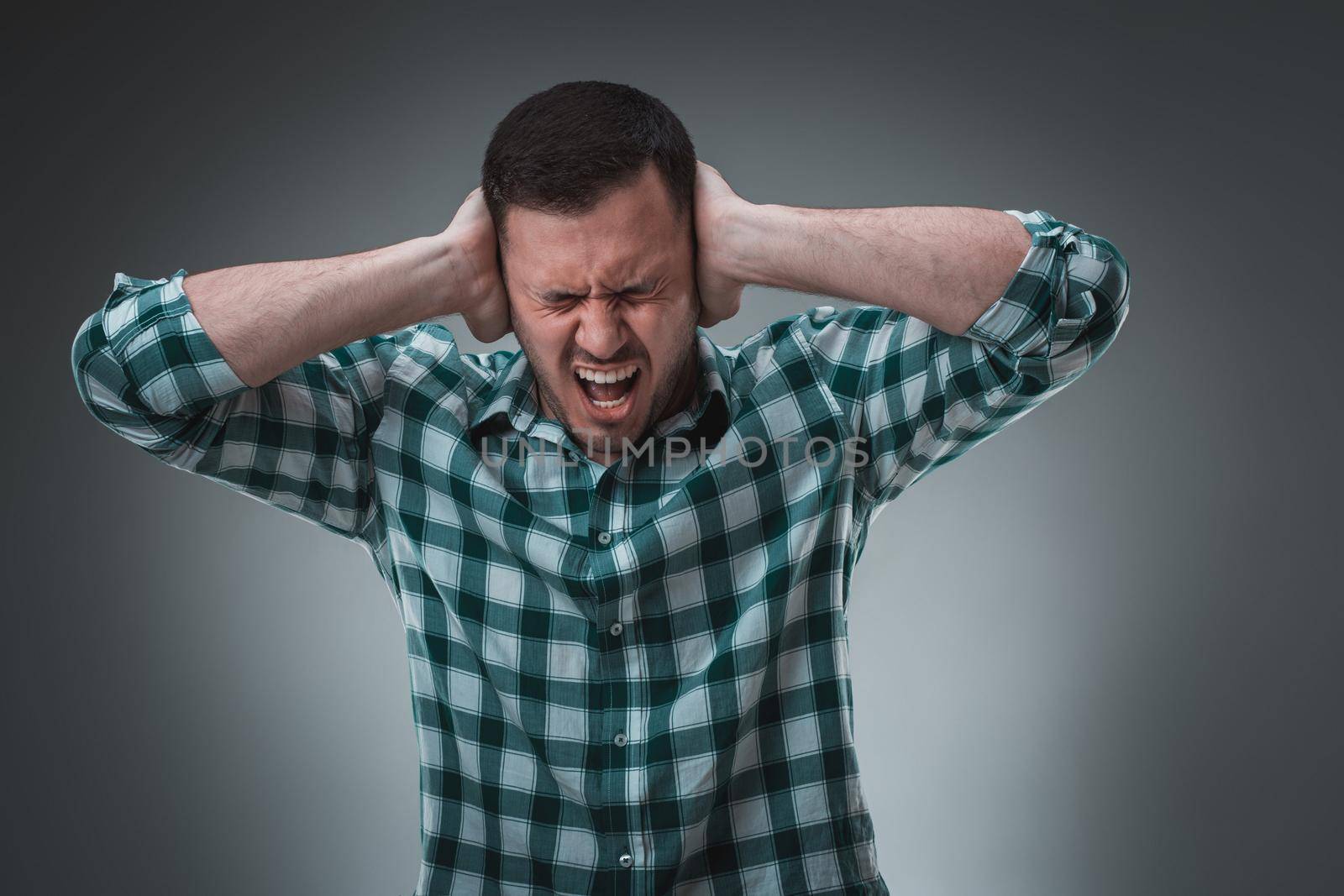 Stop that loud noise! Standing plain young man sad and depressed suffering sorrow and pain screaming desperate with hands on face against grey background. Sadness emotion concept
