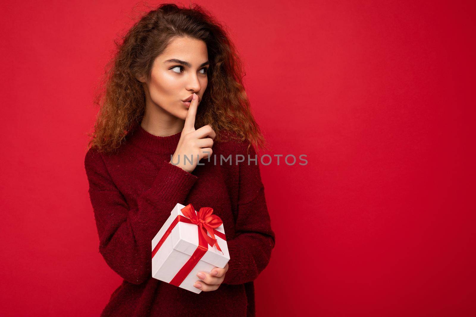 Photo shot of beautiful positive young brunette curly woman isolated over red background wall wearing red sweater holding gift box looking to the side and showing shh gesture. Copy space