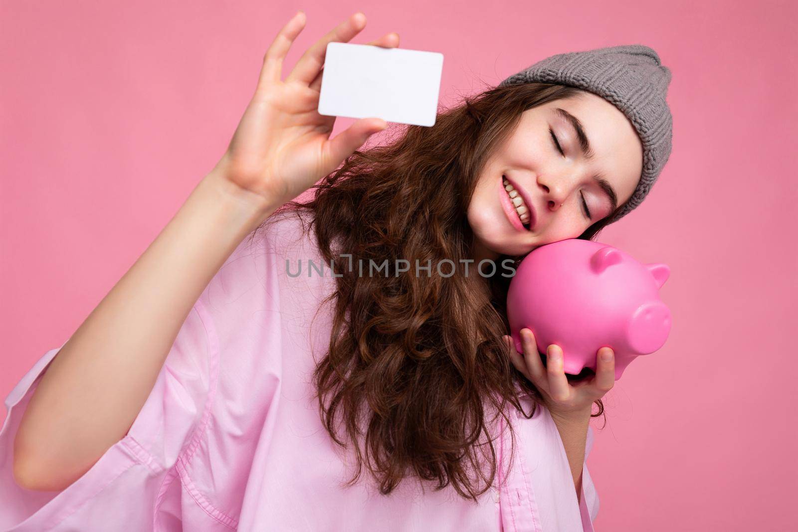 Charming smiling happy joyful young curly brunette woman wearing pink shirt and gray hat isolated on pink background with free space and holding pink pig money box and credit card for mockup.