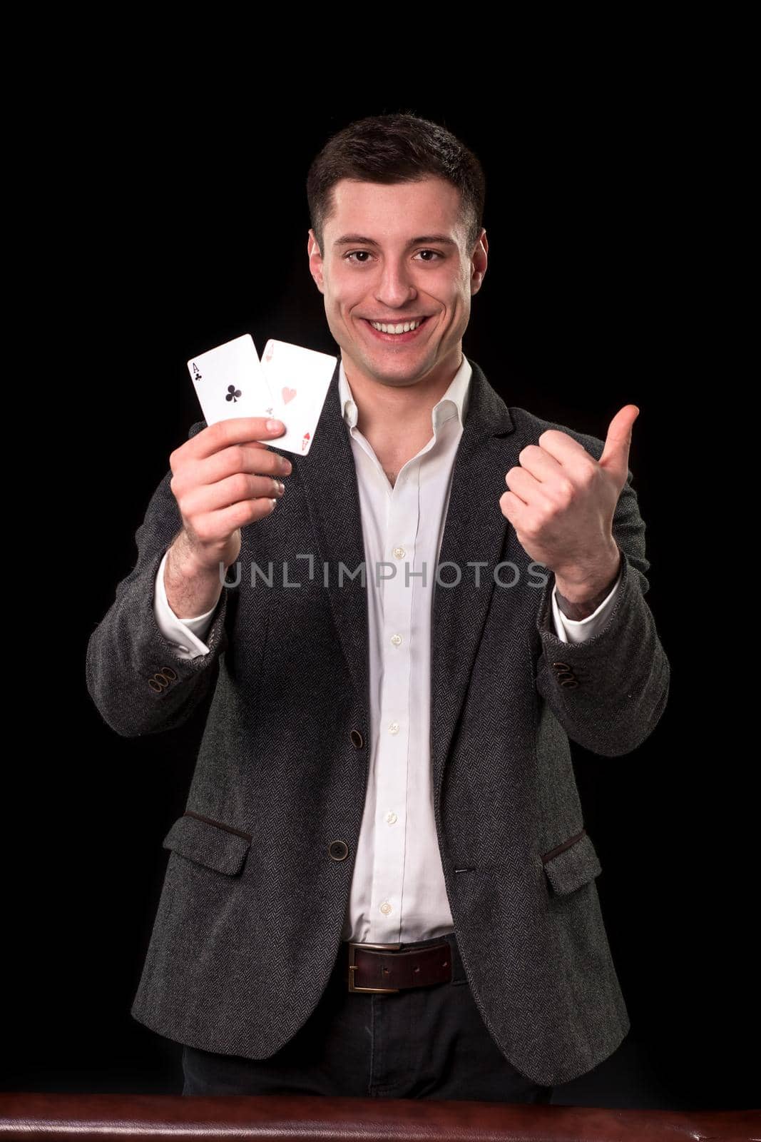 Young caucasian man wearing suit holding two aces in his hand and showing thumb up hand gesture with smile on black background. Gambling concept. Casino by nazarovsergey