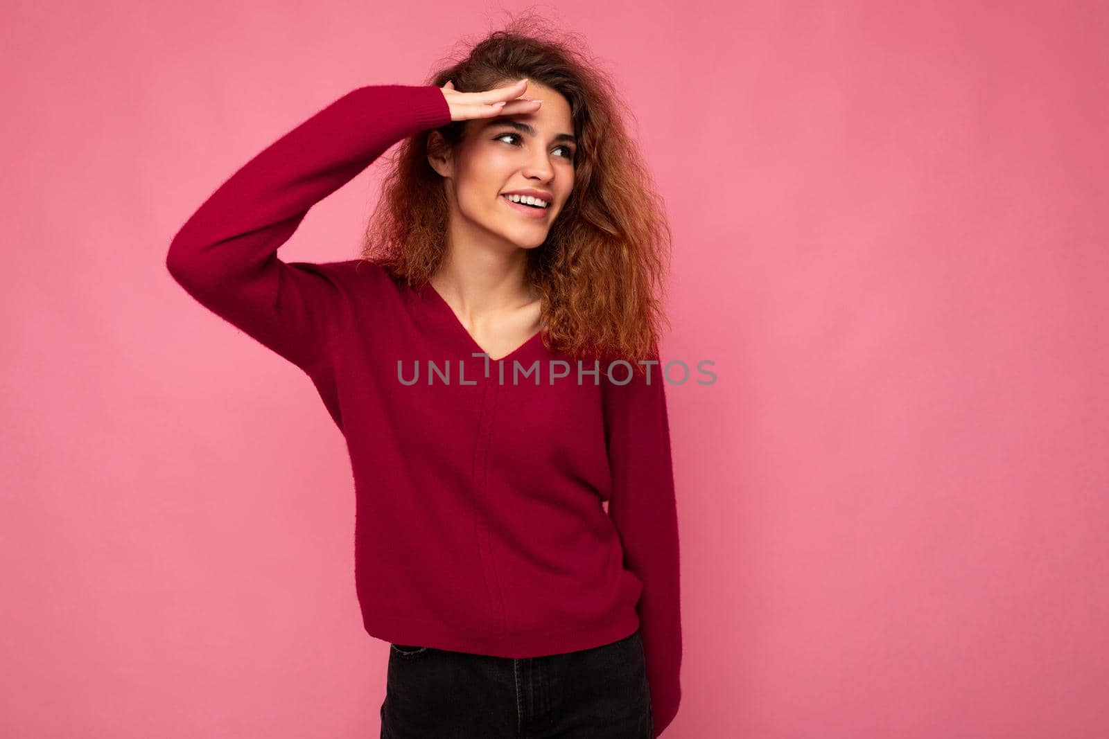 Portrait of young positive happy joyful beautiful brunette curly woman with sincere emotions wearing trendy pink pullover isolated on pink background with copy space and looking to the side.