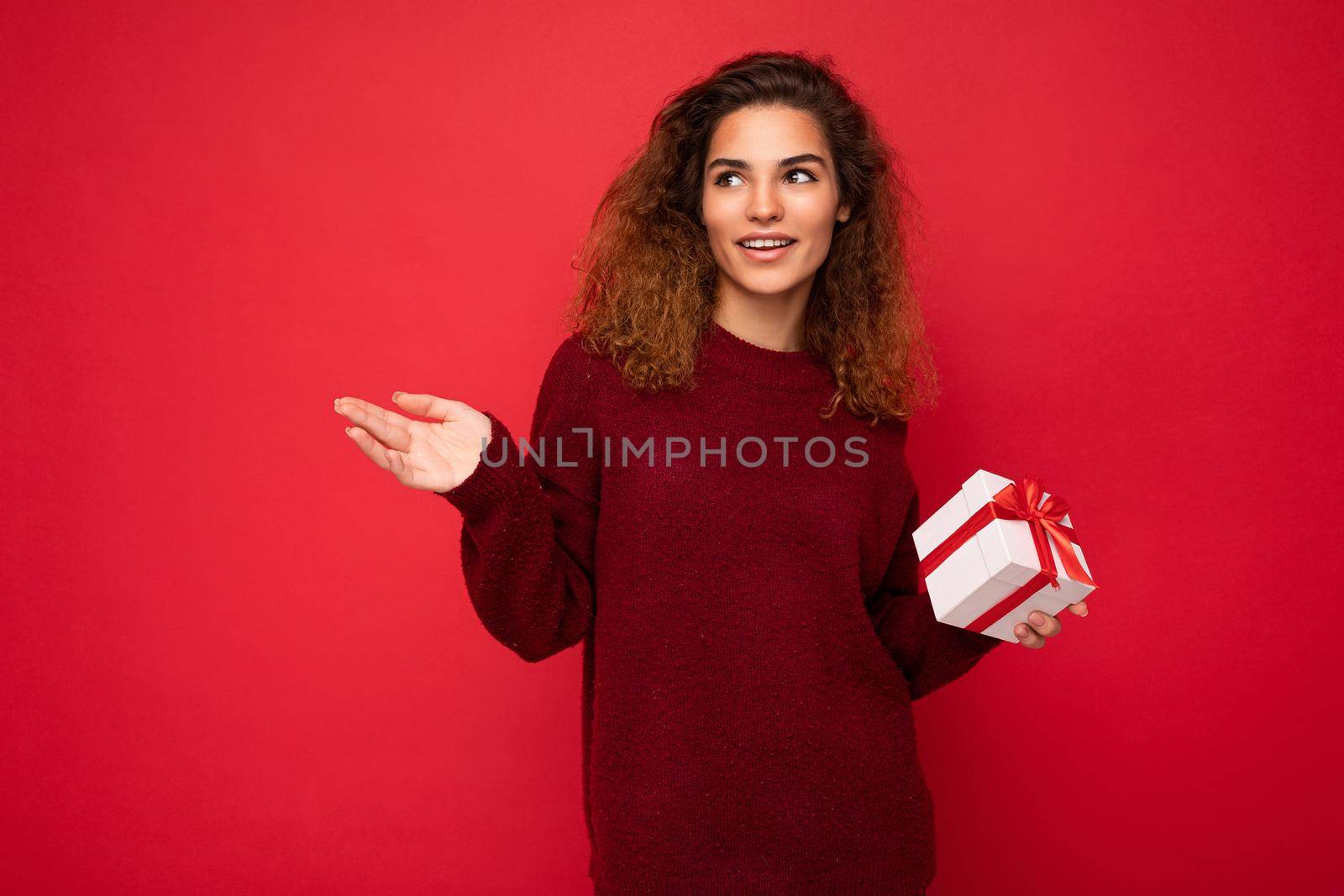 Beautiful happy young brunette woman isolated over colourful background wall wearing stylish casual clothes holding gift box and looking to the side.