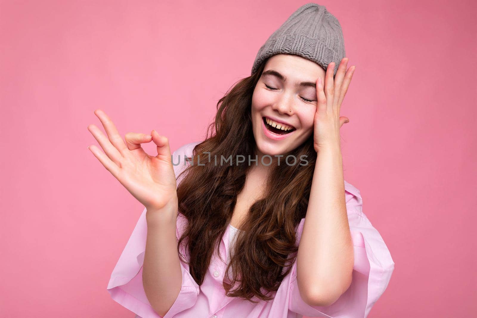 Portrait of young positive happy beautiful curly brunette woman with sincere emotions wearing trendy pink shirt and grey hat isolated on pink background with empty space and showing ok gesture. It's good.