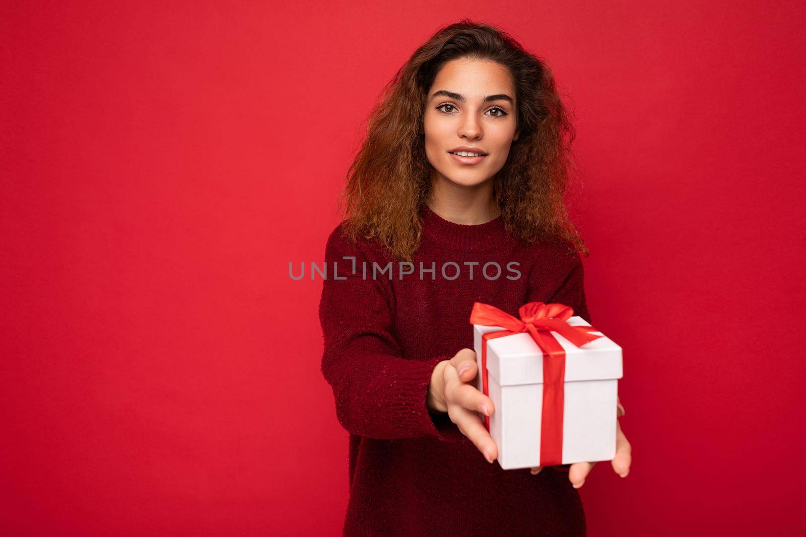 Charming happy young brunette curly woman isolated over red background wall wearing red sweater holding gift box looking at camera.