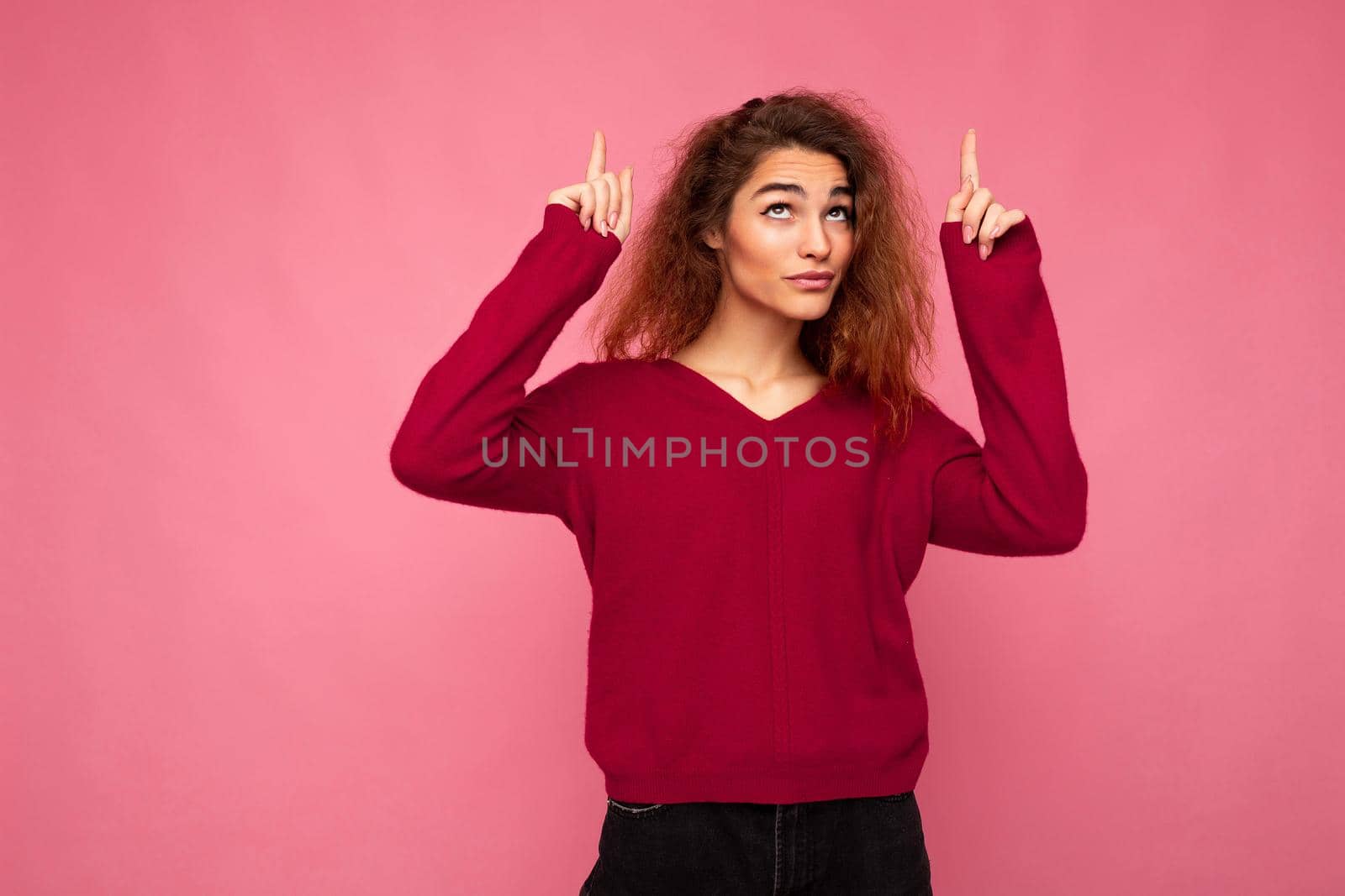 Photo of young happy positive beautiful brunette wavy woman with sincere emotions wearing casual pink jersey isolated over pink background with copy space and pointing at free space for mockup by TRMK