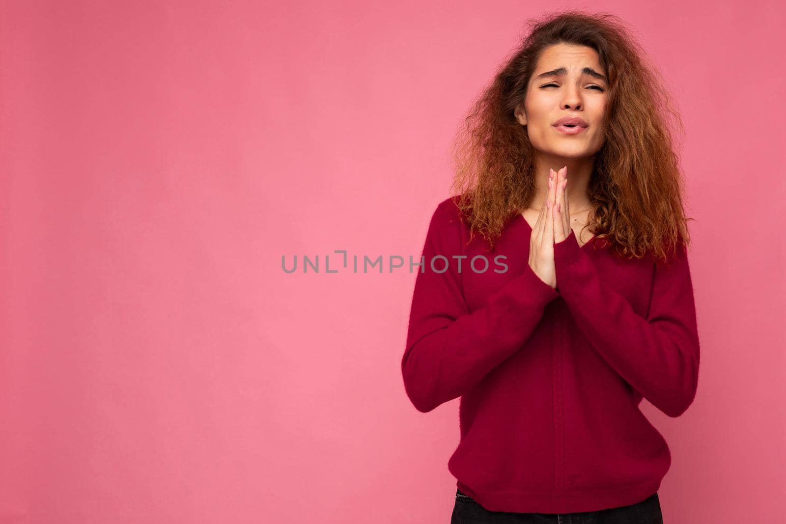 Photo of young emotional beautiful brunette curly woman with sincere emotions wearing trendy pink pullover isolated over pink background with free space and keeping hands in praying gesture and believing in good luck.