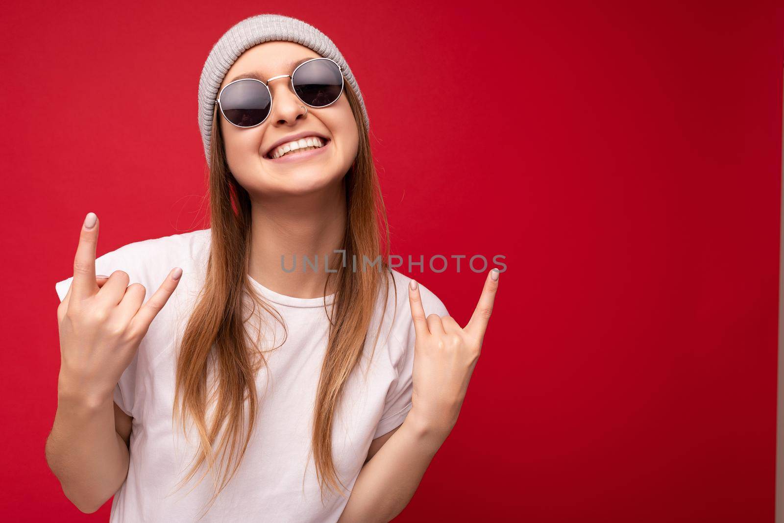 Closeup of young emotional positive happy smiling attractive dark blonde woman with sincere emotions wearing casual white t-shirt, gray hat and sunglasses isolated on red background with copy space and showing rock and roll gesture.